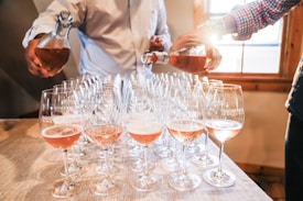 A row of wine glasses on a wooden table is being filled with rose wine by two people. The sunlight streams through a nearby window, creating a warm and inviting atmosphere. The glasses are labeled with 'Goose Gap Vineyards'.