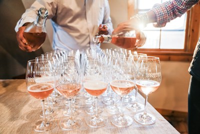 A row of wine glasses on a wooden table is being filled with rose wine by two people. The sunlight streams through a nearby window, creating a warm and inviting atmosphere. The glasses are labeled with 'Goose Gap Vineyards'.