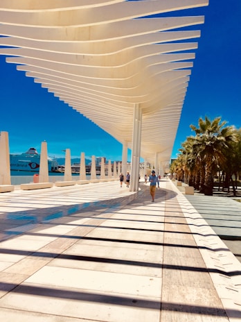 An architectural walkway with a modern canopy structure featuring wavy white beams, casting shadows on the ground. People are walking along this open, spacious port area beside a blue body of water. Palm trees line the right side, adding a touch of greenery.