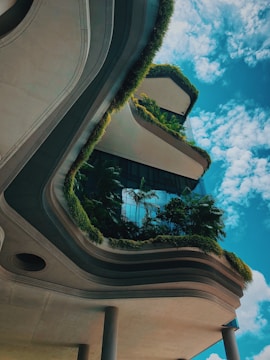 A modern architectural structure with curved lines and multiple layers, covered in lush greenery. The building features plants and foliage integrated into its design, creating a harmonious blend with nature. The sky is clear with a few clouds, adding to the serene atmosphere.