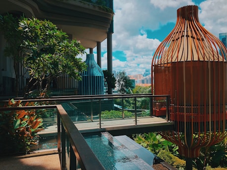 A modern architectural view featuring two large, cage-like structures with a combination of metal and wood elements. A clear blue sky with scattered clouds dominates the background, creating a bright atmosphere. Lush greenery, including trees and shrubs, surrounds a small walkway with glass railings, which passes over a tranquil water feature. Urban buildings can be seen in the distant background.