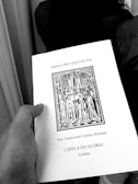 A hand holding a book titled 'MISSA PRO DEFUNCTIS' with an illustration of a religious procession, indicating a traditional Roman Catholic rite. The text also mentions 'CAPELA DA GLÓRIA' and 'Curitiba'.