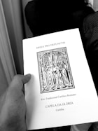 A hand holding a book titled 'MISSA PRO DEFUNCTIS' with an illustration of a religious procession, indicating a traditional Roman Catholic rite. The text also mentions 'CAPELA DA GLÓRIA' and 'Curitiba'.