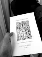 A hand holding a book titled 'MISSA PRO DEFUNCTIS' with an illustration of a religious procession, indicating a traditional Roman Catholic rite. The text also mentions 'CAPELA DA GLÓRIA' and 'Curitiba'.