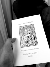 A hand holding a book titled 'MISSA PRO DEFUNCTIS' with an illustration of a religious procession, indicating a traditional Roman Catholic rite. The text also mentions 'CAPELA DA GLÓRIA' and 'Curitiba'.