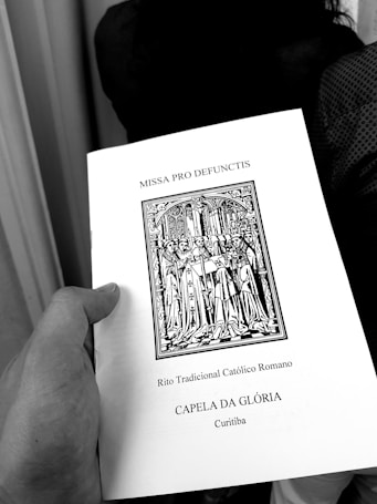 A hand holding a book titled 'MISSA PRO DEFUNCTIS' with an illustration of a religious procession, indicating a traditional Roman Catholic rite. The text also mentions 'CAPELA DA GLÓRIA' and 'Curitiba'.