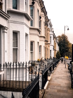 A row of elegant Victorian-style townhouses lines a quiet street. The buildings feature a mix of white and beige bricks with elaborate iron railings along the front steps. The street is bordered by a narrow sidewalk with a few lampposts. Trees can be seen in the distance, and a couple of people walk along the pavement.