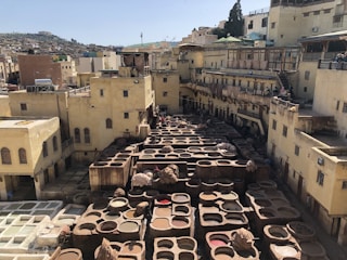 A traditional tannery with numerous stone vats filled with various colored dyes used in leather processing. Surrounding the vats are historical buildings made of beige-colored stone or mud, typical of North African architecture. People are seen working or walking around the area. Rooftops and distant buildings form the background, giving a sense of a busy urban environment.