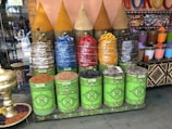 A vibrant display of colorful spices and stones in decorative green cans, topped with conical covers. The spices are arranged in a market-like setting with a variety of colors including yellow, red, blue, and black. Nearby, a selection of patterned cones contains more pigments. A large ornate gold bowl is placed on the floor filled with dried ingredients.