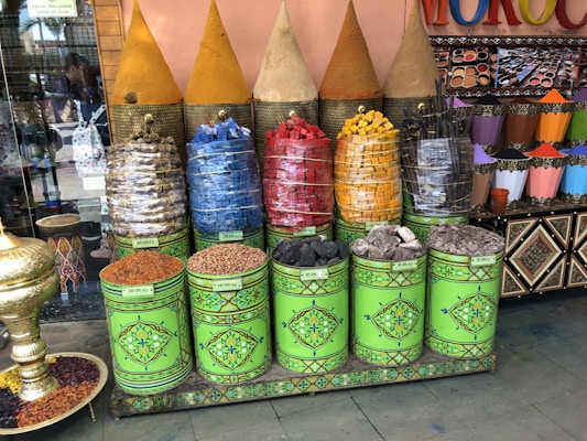 A vibrant display of colorful spices and stones in decorative green cans, topped with conical covers. The spices are arranged in a market-like setting with a variety of colors including yellow, red, blue, and black. Nearby, a selection of patterned cones contains more pigments. A large ornate gold bowl is placed on the floor filled with dried ingredients.