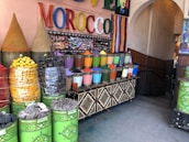 A market stall displaying a vibrant array of colored powders, likely pigments or spices, arranged in neat metal containers. The setting includes large cones of products, stacked baskets of colorful items, and a sign with the word MOROCCO in bright, multicolored letters. The area is decorated with intricate patterns and colorful stripes.