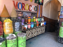 A market stall displaying a vibrant array of colored powders, likely pigments or spices, arranged in neat metal containers. The setting includes large cones of products, stacked baskets of colorful items, and a sign with the word MOROCCO in bright, multicolored letters. The area is decorated with intricate patterns and colorful stripes.