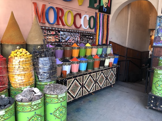 A market stall displaying a vibrant array of colored powders, likely pigments or spices, arranged in neat metal containers. The setting includes large cones of products, stacked baskets of colorful items, and a sign with the word MOROCCO in bright, multicolored letters. The area is decorated with intricate patterns and colorful stripes.