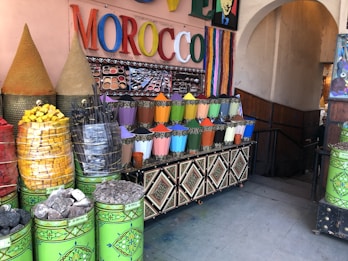 A market stall displaying a vibrant array of colored powders, likely pigments or spices, arranged in neat metal containers. The setting includes large cones of products, stacked baskets of colorful items, and a sign with the word MOROCCO in bright, multicolored letters. The area is decorated with intricate patterns and colorful stripes.