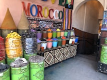 A market stall displaying a vibrant array of colored powders, likely pigments or spices, arranged in neat metal containers. The setting includes large cones of products, stacked baskets of colorful items, and a sign with the word MOROCCO in bright, multicolored letters. The area is decorated with intricate patterns and colorful stripes.