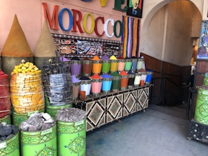 A market stall displaying a vibrant array of colored powders, likely pigments or spices, arranged in neat metal containers. The setting includes large cones of products, stacked baskets of colorful items, and a sign with the word MOROCCO in bright, multicolored letters. The area is decorated with intricate patterns and colorful stripes.