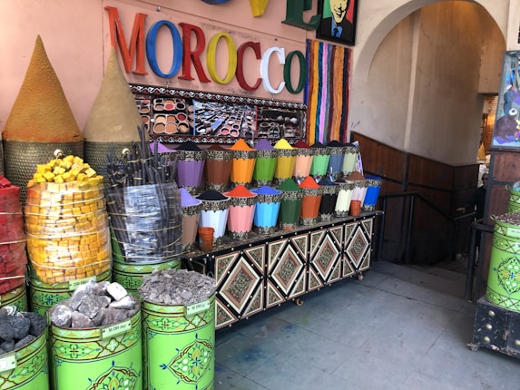 A market stall displaying a vibrant array of colored powders, likely pigments or spices, arranged in neat metal containers. The setting includes large cones of products, stacked baskets of colorful items, and a sign with the word MOROCCO in bright, multicolored letters. The area is decorated with intricate patterns and colorful stripes.