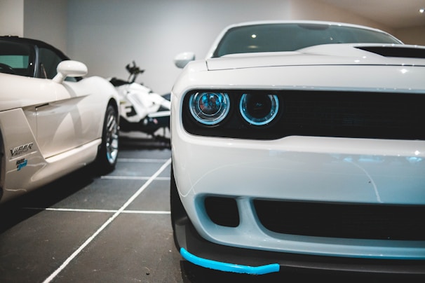 A close-up view of a sleek white sports car with a prominent front grille and distinctive headlights. Adjacent to this car, another white car can be seen partially, and a motorcycle is in the background, adding to the high-performance vehicles theme.