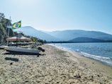 A peaceful beach scene with a small boat on the sand, featuring a Brazilian flag. The shoreline extends into the distance, bordered by gentle waves from the ocean. Mountains provide a scenic backdrop, and the sky is clear with a bright blue hue.