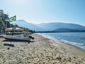 A peaceful beach scene with a small boat on the sand, featuring a Brazilian flag. The shoreline extends into the distance, bordered by gentle waves from the ocean. Mountains provide a scenic backdrop, and the sky is clear with a bright blue hue.