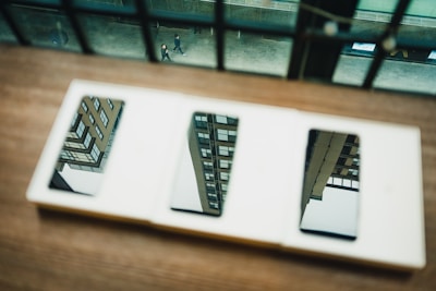 A set of mirrors on a wooden surface reflecting the image of modern buildings. In the background, a street view with people walking can be seen through a window with vertical bars.