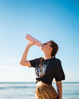 woman drinking water using pink bottle beside sea during daytime