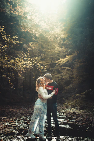 A boyfriend and girlfriend smiling warmly at each other in a sunlit forest clearing.