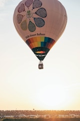 A hot air balloon adorned with a colorful flower and panels featuring text is soaring through the sky. The scene is set against a clear sky during sunset, creating a warm, golden atmosphere. Below, a large crowd of people and several vehicles are visible on the ground, silhouetted against the bright sunlight.