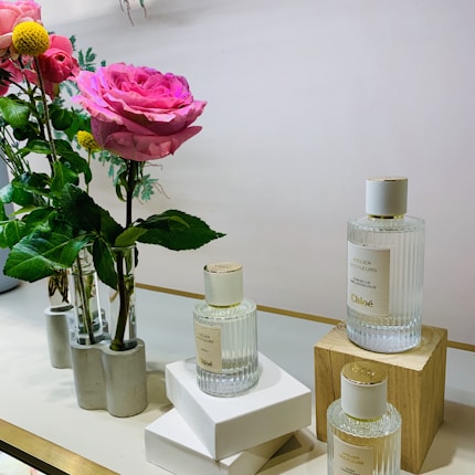 On a light-colored surface, three elegant glass perfume bottles are displayed next to vases holding freshly cut flowers, including large pink roses and green leaves. The bottles have a simple, luxurious design, and one is elevated on a wooden block.