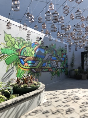 An artistic space features a vibrant mural on the wall with lush greenery and abstract shapes. Numerous decorative birdcages hang from the ceiling in a symmetrical pattern, casting shadows on the floor. The setting includes potted plants, adding to the natural theme.