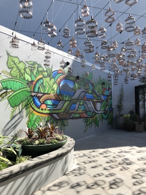 An artistic space features a vibrant mural on the wall with lush greenery and abstract shapes. Numerous decorative birdcages hang from the ceiling in a symmetrical pattern, casting shadows on the floor. The setting includes potted plants, adding to the natural theme.