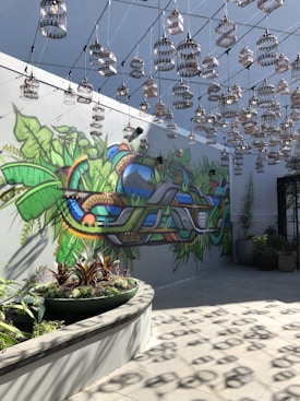 An artistic space features a vibrant mural on the wall with lush greenery and abstract shapes. Numerous decorative birdcages hang from the ceiling in a symmetrical pattern, casting shadows on the floor. The setting includes potted plants, adding to the natural theme.