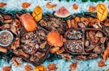 A vibrant and colorful seafood feast is displayed, featuring large crabs, shrimp, and mussels surrounded by rice and garnished with sliced mangoes and watermelon. Various grilled meats and a dish of dipping sauce are arranged on wooden platters, creating an inviting and appetizing arrangement.