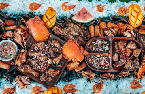 A vibrant and colorful seafood feast is displayed, featuring large crabs, shrimp, and mussels surrounded by rice and garnished with sliced mangoes and watermelon. Various grilled meats and a dish of dipping sauce are arranged on wooden platters, creating an inviting and appetizing arrangement.