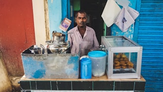 A man stands behind a small street food stall that has a variety of utensils and containers. The stall includes a steel container with a spout, a blue plastic container, and a glass display case with fried snacks. The background features a combination of blue and red walls, with newspapers and bags pinned to the wall.