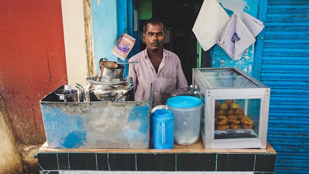 A man stands behind a small street food stall that has a variety of utensils and containers. The stall includes a steel container with a spout, a blue plastic container, and a glass display case with fried snacks. The background features a combination of blue and red walls, with newspapers and bags pinned to the wall.