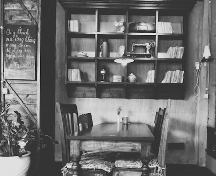 A cozy and rustic interior featuring a wooden table with two chairs, an open shelf filled with old books and vintage items like a fan and sewing machine. The wall displays a chalkboard with some writing, and a potted plant adds a touch of greenery. The space feels intimate and quaint.