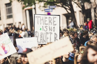 A group of people participating in a protest or march, holding signs. One prominent sign in the foreground reads 'There is no Planet B.' The scene takes place outdoors, with buildings and trees in the background.