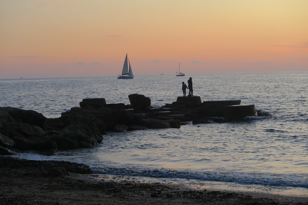 two people standing boulder near seashore during sunset