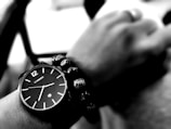A black and white close-up of a wrist wearing a watch and a bracelet. The watch has a minimalist design with a dark face and white hour markers. The bracelet consists of round beads with a patterned texture, adding a contrast to the smooth watch surface. The background is blurred, highlighting the wrist and accessories.