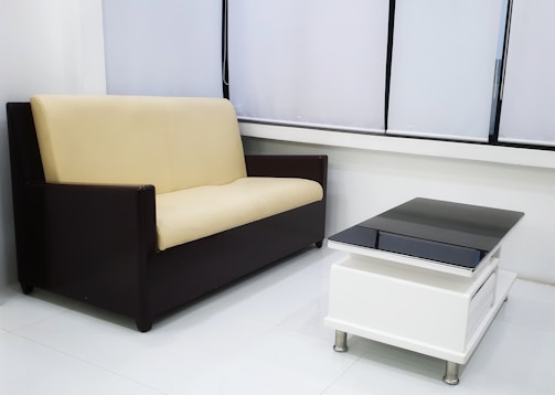 A modern living room setup featuring a beige and black sofa placed along a wall with large windows covered by white blinds. In front of the sofa, a sleek black and white coffee table with metal legs sits on a polished white floor.