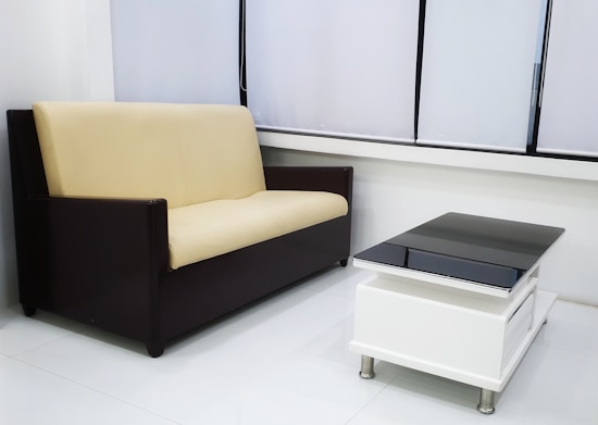 A modern living room setup featuring a beige and black sofa placed along a wall with large windows covered by white blinds. In front of the sofa, a sleek black and white coffee table with metal legs sits on a polished white floor.
