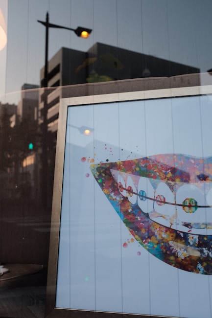 A framed artwork displays a stylized, colorful depiction of a smiling mouth with braces. The background is white, and the artwork features vibrant splashes of color, including reds, blues, greens, and yellows, forming an abstract representation. A glass reflection shows a street scene with buildings and streetlights.
