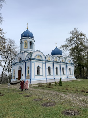 A beautiful Orthodox church with blue domes and white walls is situated amidst a lush green lawn. The building features ornate decorative elements and icons on its exterior. Two people are walking along a stone pathway leading to the entrance of the church. Tall trees frame the background, and the sky is overcast.