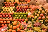 Various types of fresh fruits are neatly arranged in stacks. The display includes apples, pears, grapes, oranges, pomegranates, and melons. Some pumpkins and gourds are also present. The fruits are vibrant in color and appear to be in excellent condition. Labels are visible on some of the fruits, and there are also some informational packets labeled 'Sweet Tamarind'. The background includes some decorative elements, adding to the freshness and appeal of the display.