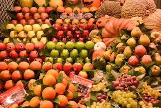 Various types of fresh fruits are neatly arranged in stacks. The display includes apples, pears, grapes, oranges, pomegranates, and melons. Some pumpkins and gourds are also present. The fruits are vibrant in color and appear to be in excellent condition. Labels are visible on some of the fruits, and there are also some informational packets labeled 'Sweet Tamarind'. The background includes some decorative elements, adding to the freshness and appeal of the display.