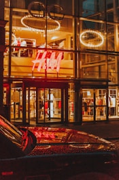 A brightly illuminated storefront with a large H&M logo, featuring clothing displays visible through the glass facade. Circular light fixtures are seen hanging from the ceiling inside the store. The reflective surface of a car is visible in the foreground, capturing the store lights.