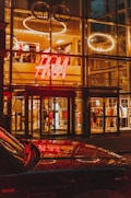 A brightly illuminated storefront with a large H&M logo, featuring clothing displays visible through the glass facade. Circular light fixtures are seen hanging from the ceiling inside the store. The reflective surface of a car is visible in the foreground, capturing the store lights.
