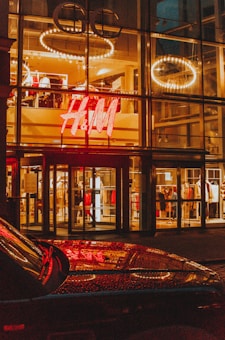 A brightly illuminated storefront with a large H&M logo, featuring clothing displays visible through the glass facade. Circular light fixtures are seen hanging from the ceiling inside the store. The reflective surface of a car is visible in the foreground, capturing the store lights.