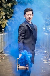 A man in a black coat holds a device releasing blue smoke, surrounded by foliage and metal railings in an outdoor setting.